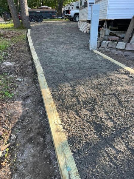 Gravel pathway with wooden edging leading to a camping trailer in a wooded area.