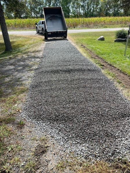 Dump truck unloading gravel for a new driveway beside green grass and trees.
