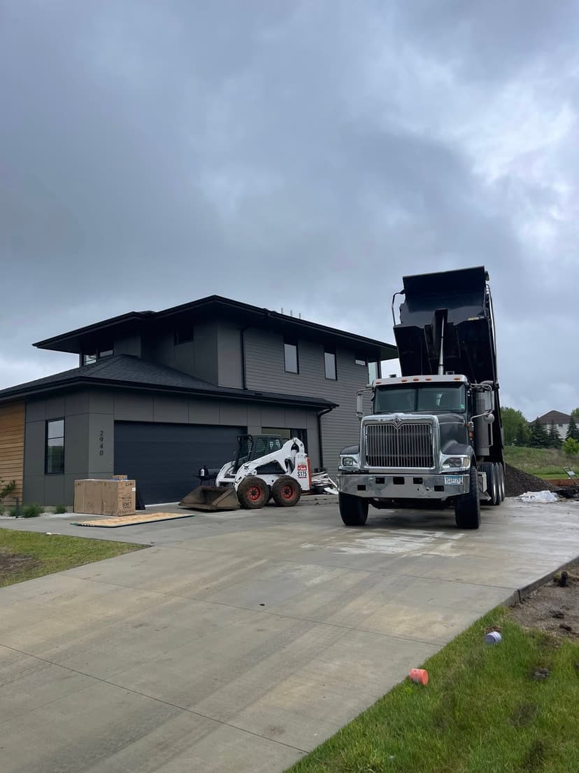 Dump truck unloading material near modern home with construction equipment on driveway.