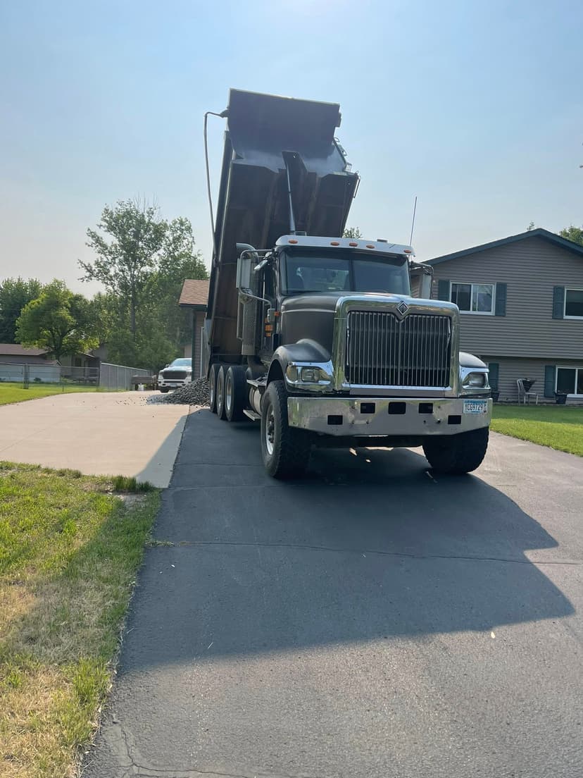 Dump truck unloading materials on a residential driveway with a house in the background.
