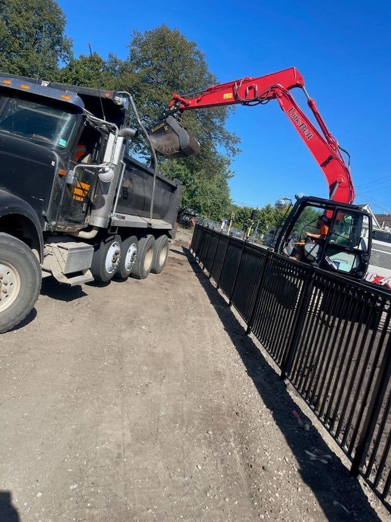 Excavator loading material from a dump truck by a fence on a construction site.