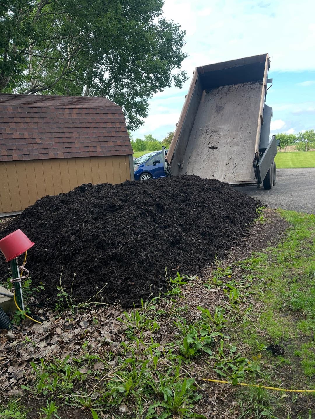 Truck unloading a large pile of dark mulch beside a storage shed on a sunny day.