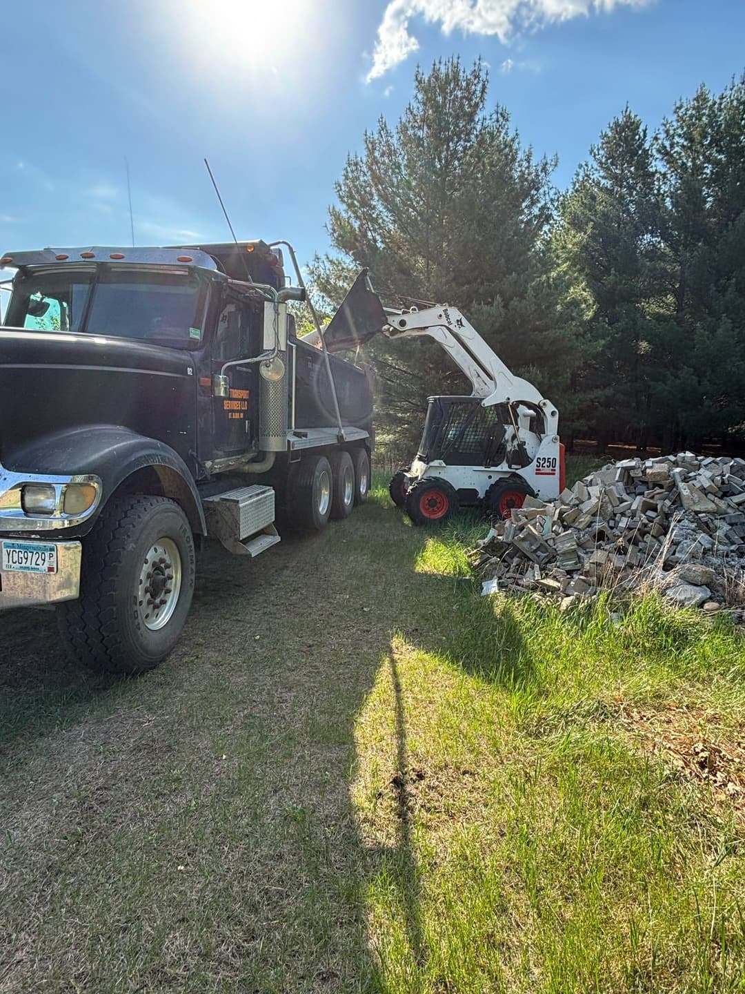 Dump truck and skid steer loader in a forested area with a pile of rocks nearby.