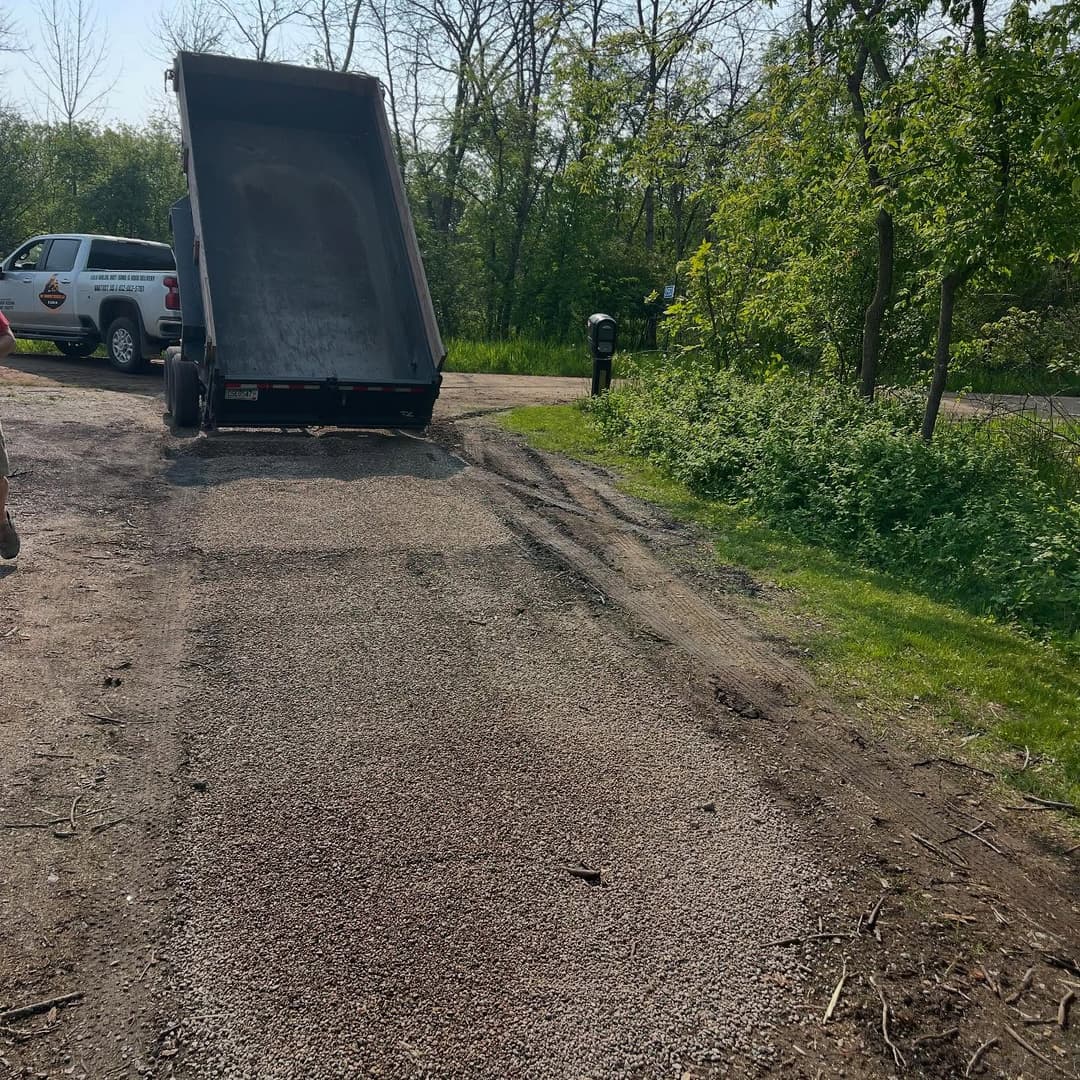 Dump truck unloading gravel on a gravel road near a mailbox and green foliage.