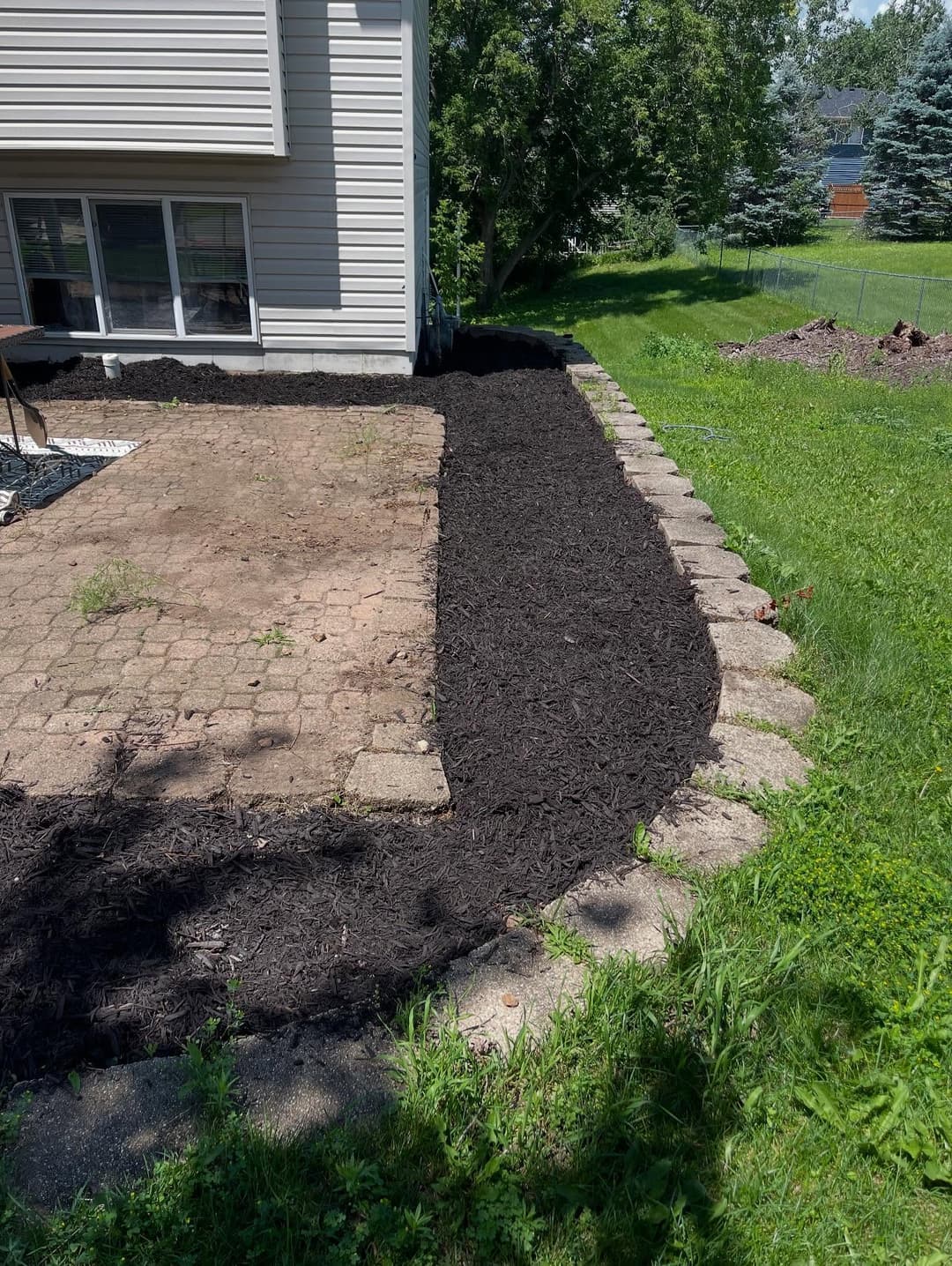 Freshly laid black mulch along a paved garden path beside a house. Green grass and trees in background.