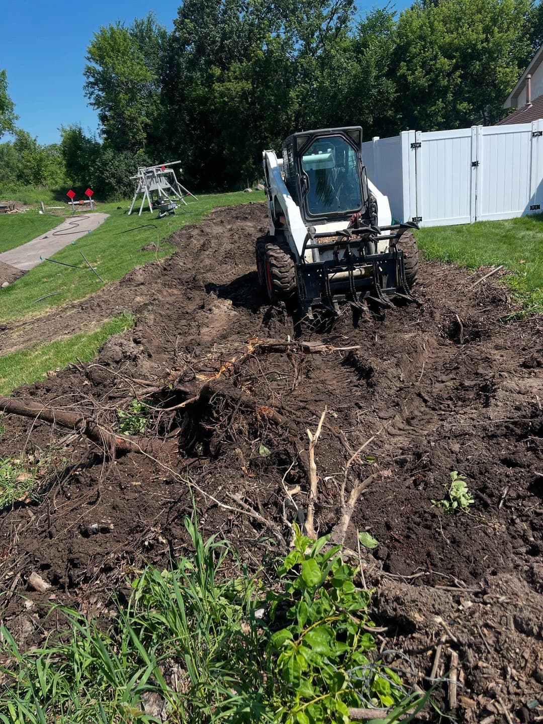 Construction site with a Bobcat skid steer clearing land, surrounded by greenery and a fence.
