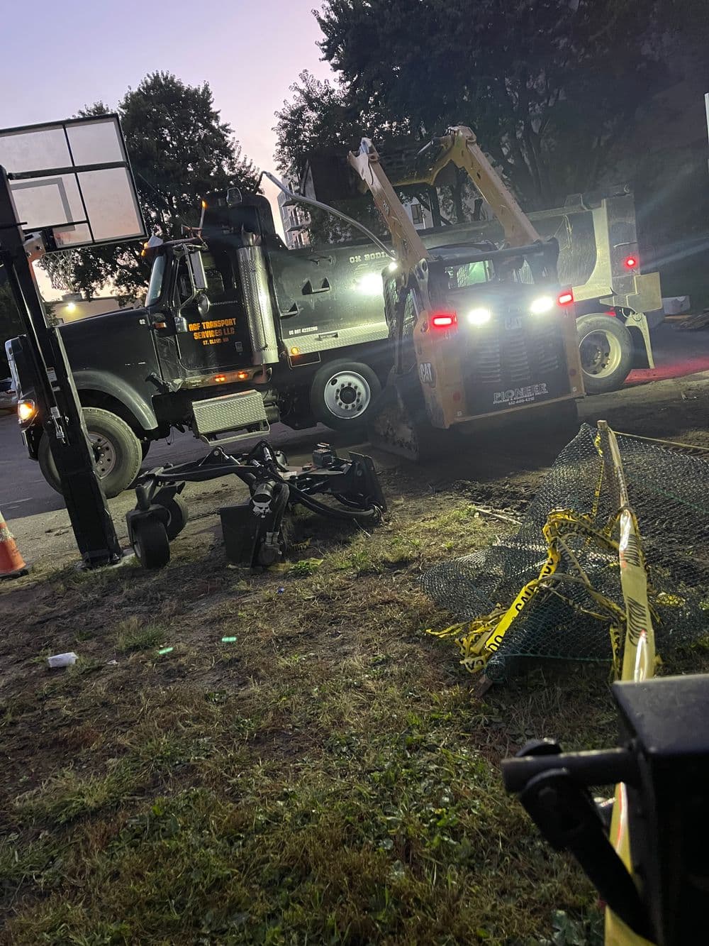 Construction vehicles working near a basketball court at dusk, with safety equipment visible.
