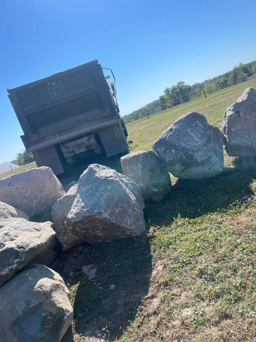 Dump truck unloading large rocks on a grassy landscape under a clear blue sky.