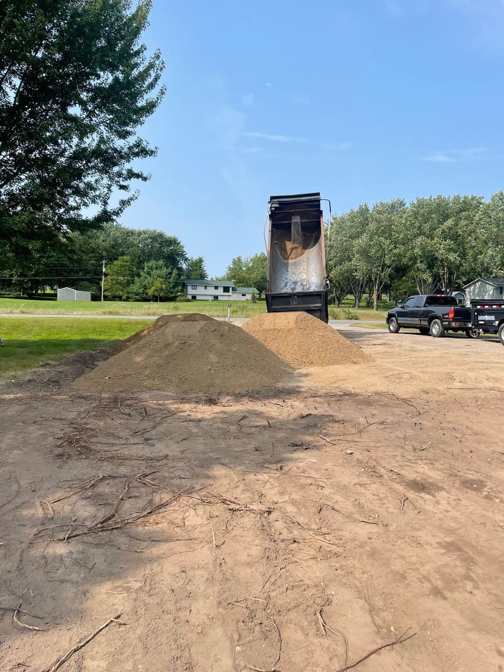 Dump truck unloading sand at construction site with trees and parked vehicles in background.