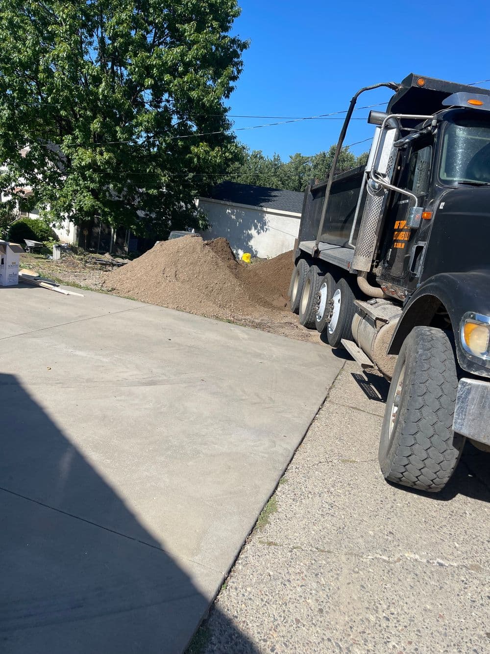 Dump truck unloading a large pile of dirt in a residential driveway under a clear blue sky.
