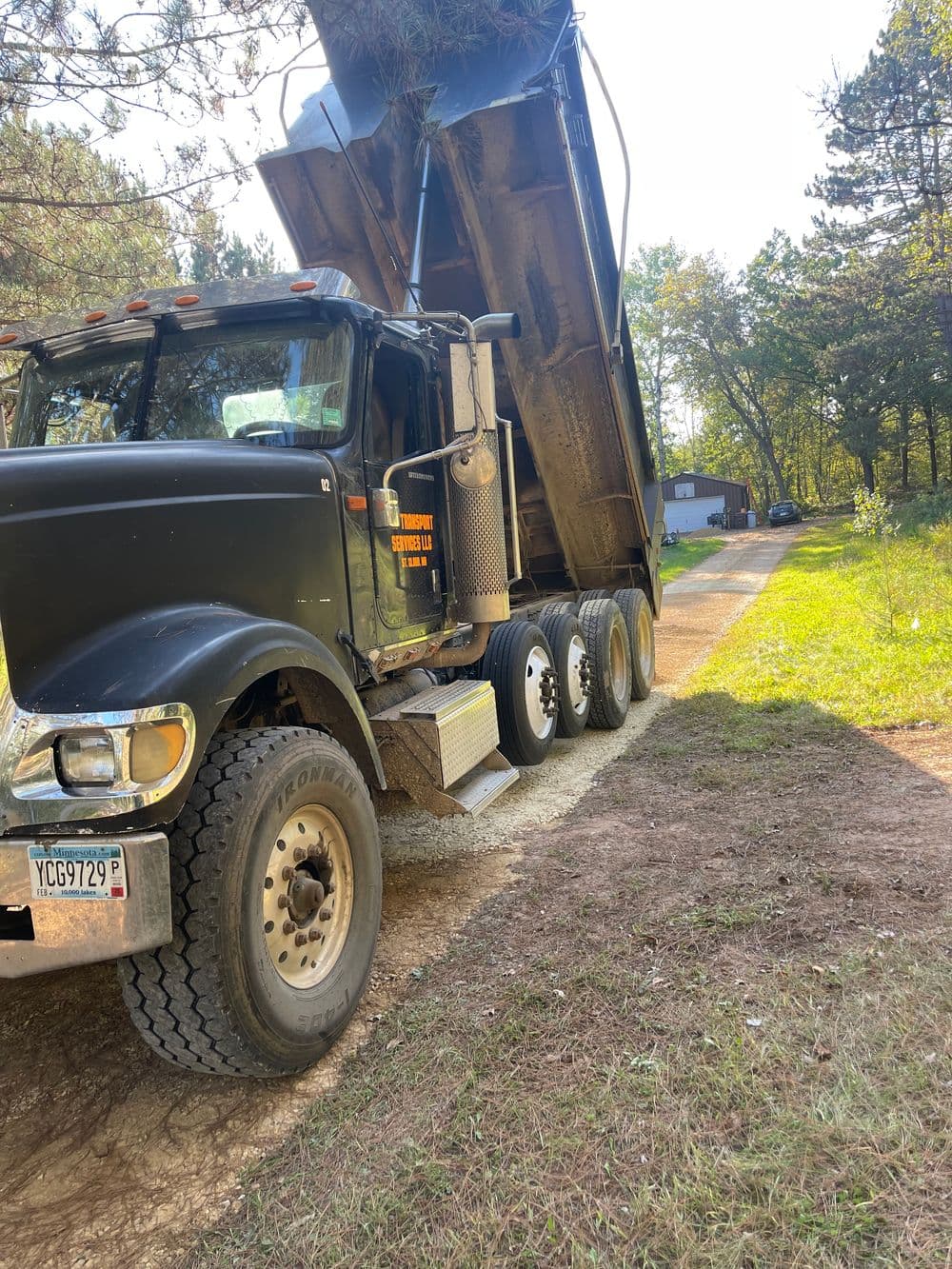 Dump truck unloading gravel on a gravel driveway surrounded by trees.