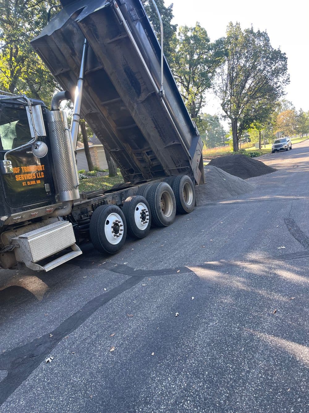 Dump truck unloading gravel on a residential street with trees in the background.