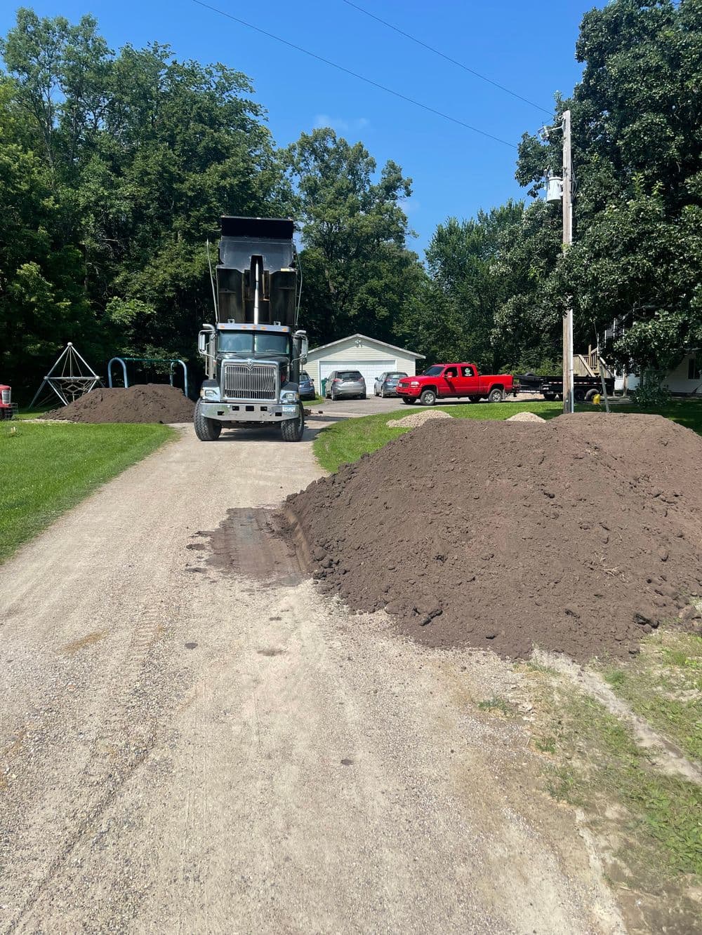 Dump truck unloading dirt on a gravel driveway with piles of soil and parked vehicles nearby.