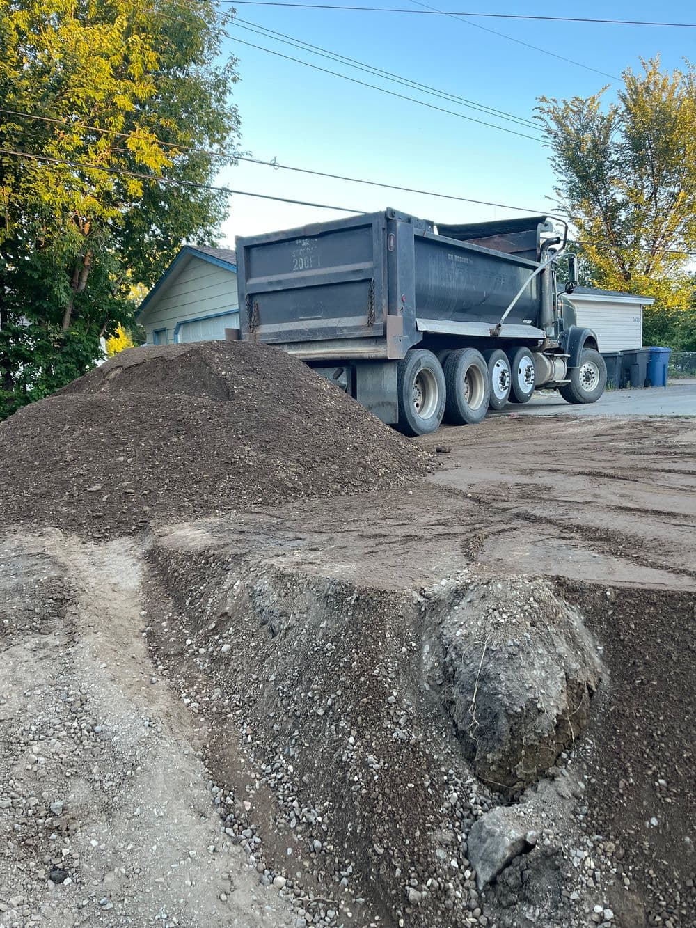Dump truck unloading soil at a residential site with a gravel surface and trees in the background.