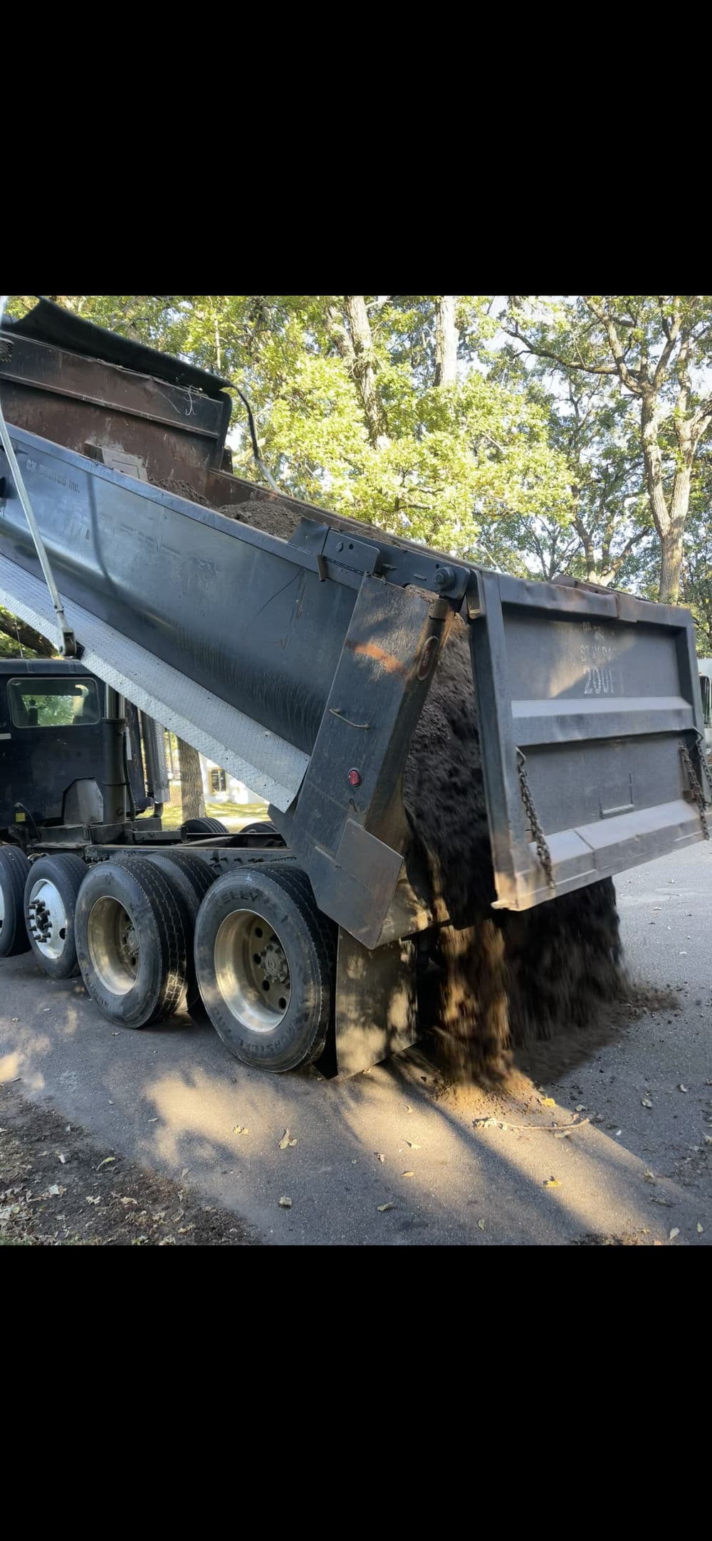 Dump truck unloading dirt on a residential street surrounded by trees.