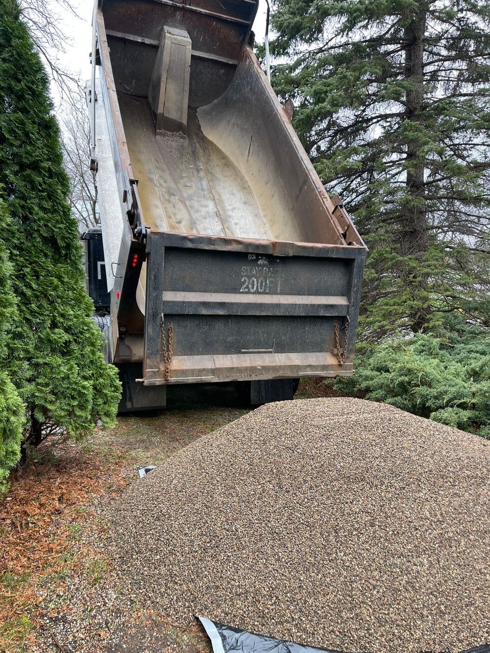 Dump truck unloading gravel onto a landscaped area with trees in the background.