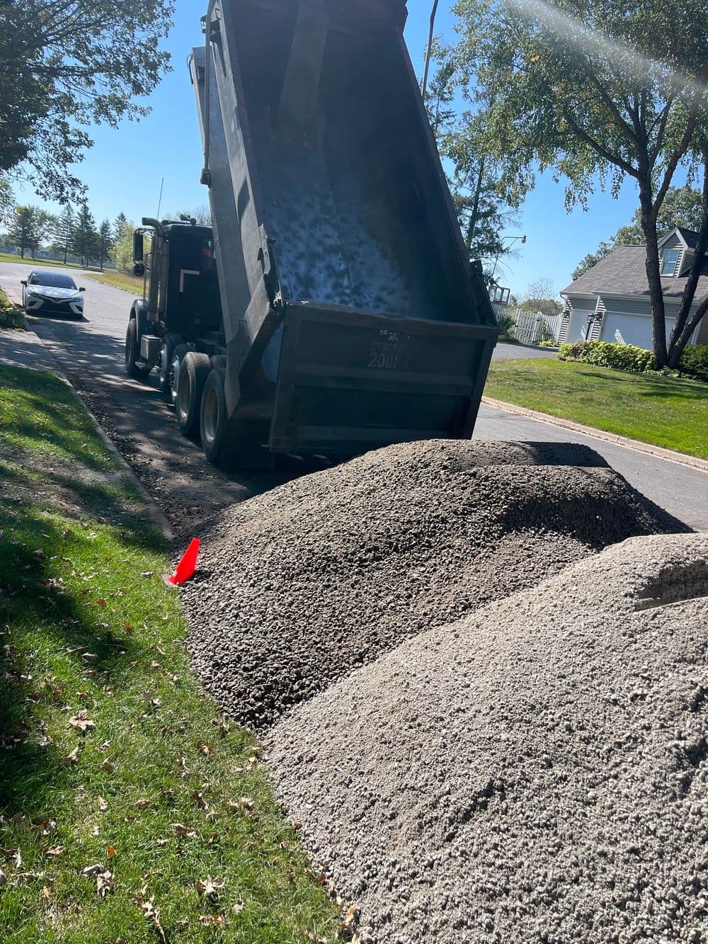 Dump truck unloading gravel onto a residential street, with piles of gravel nearby.